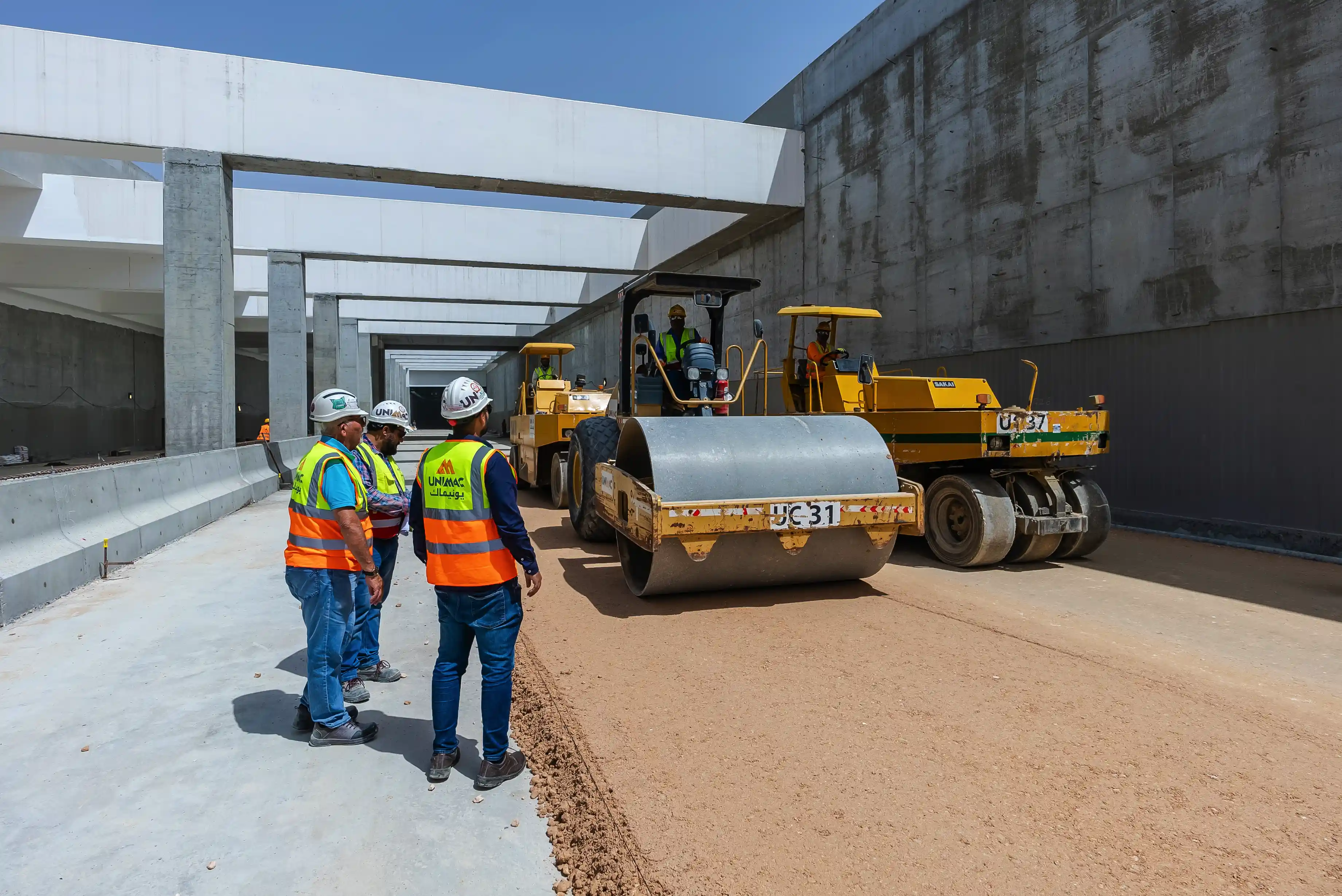 Construction workers in safety vests and helmets observing two yellow road rollers compacting soil inside a concrete structure.