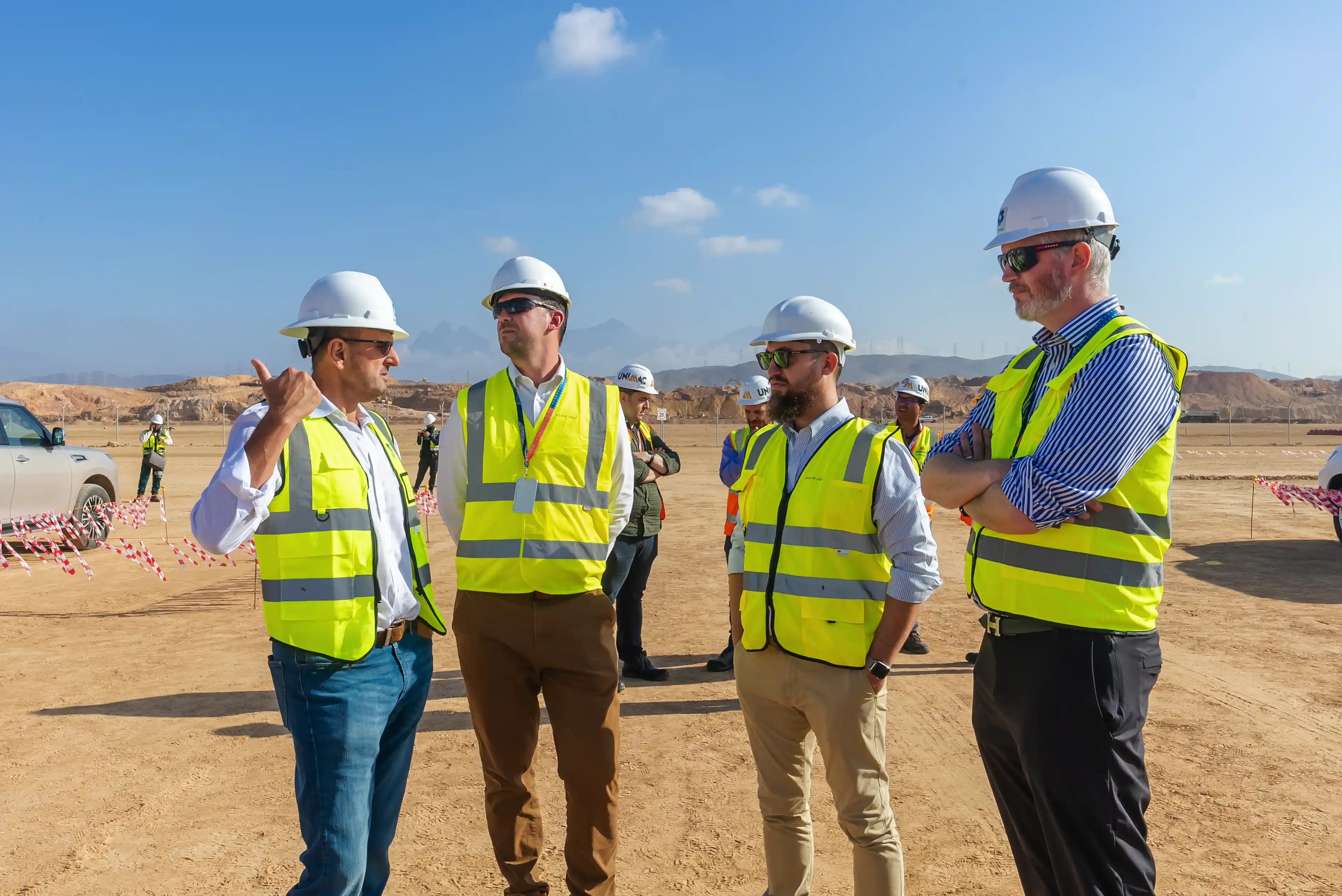 Four men wearing white hard hats, sunglasses, and yellow safety vests stand on a construction site having a discussion.