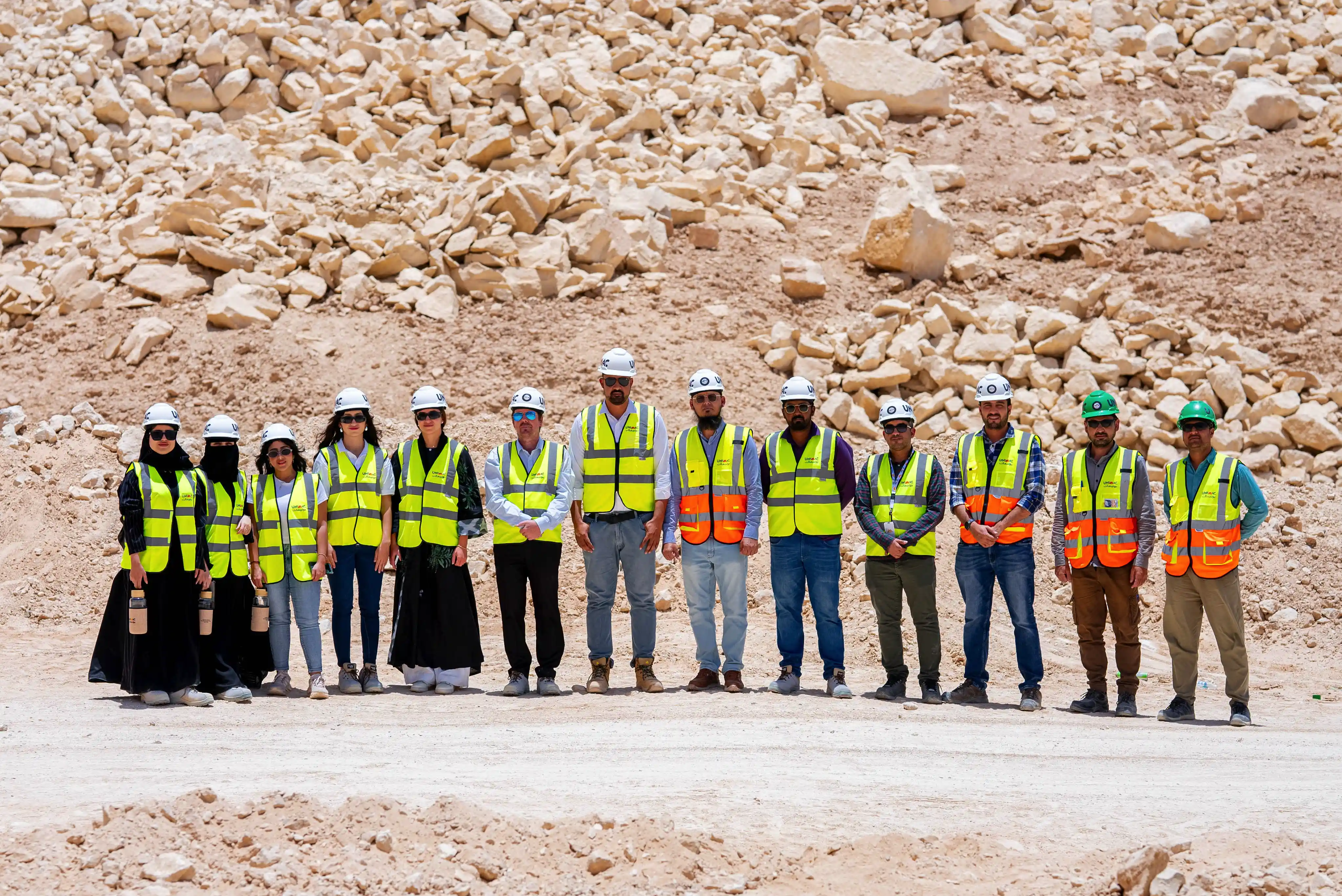 A group of fourteen construction workers wearing safety vests and helmets standing in a rocky outdoor setting.